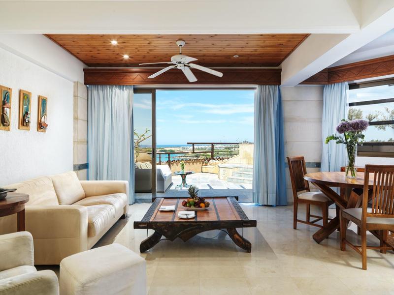 Helles Wohnzimmer mit beiger Couch, Holztisch, Esstisch, Blumen und Meerblick durch Glasschiebetüren.