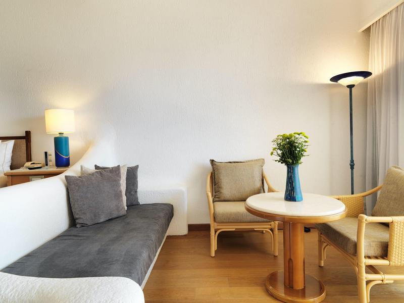 Wohnzimmer mit grauem Sofa, zwei Holzsesseln, runden Holztisch und Blumen in blauer Vase.