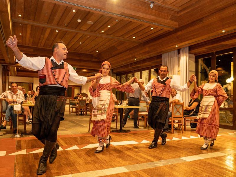 Vier Menschen tanzen in traditioneller Tracht in einem warm beleuchteten Saal mit Holzboden.
