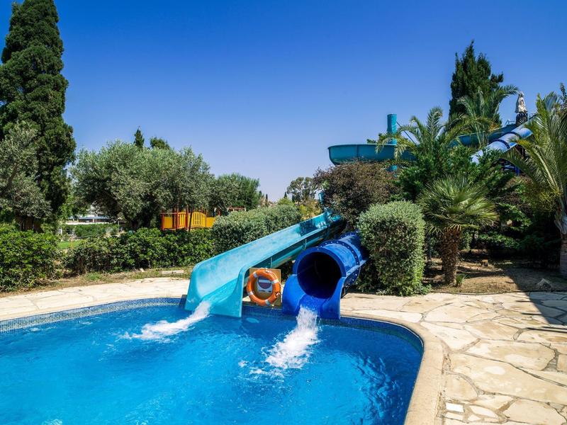 Pool mit zwei blauen Wasserrutschen inmitten von Palmen und Grünpflanzen an sonnigem Tag.