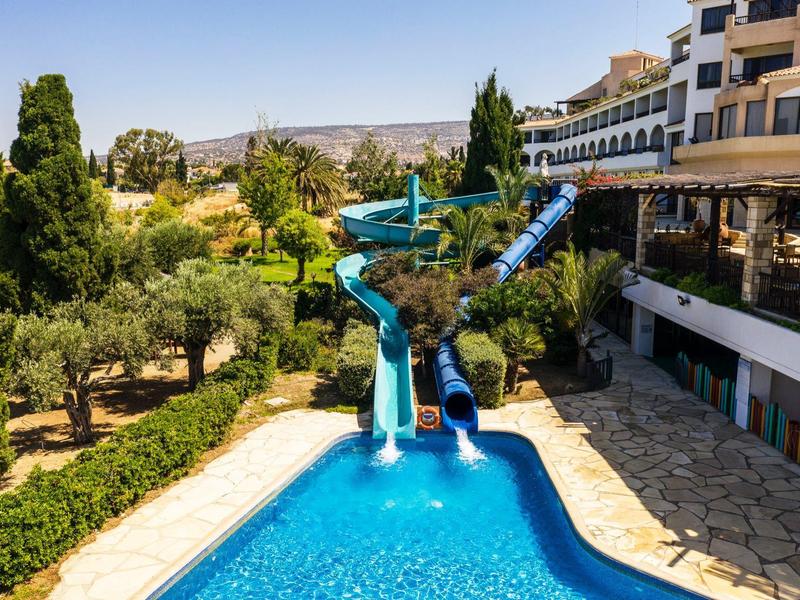 Blaue Wasserrutschen führen in ein Schwimmbecken, umgeben von Bäumen und einem mehrstöckigen Gebäude.