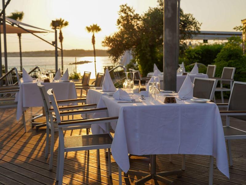 Terrasse mit gedeckten Tischen und Stühlen, Sonnenuntergang über Wasser und Palmen im Hintergrund.