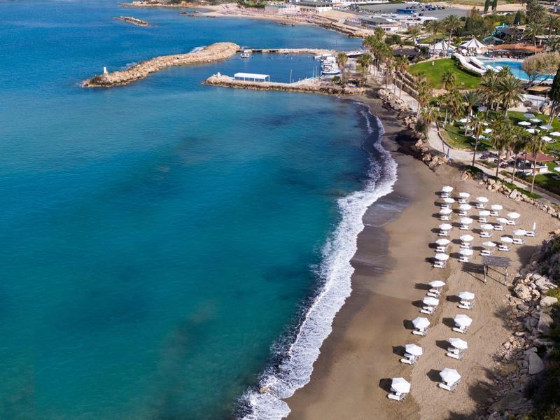 Luftaufnahme eines Sandstrands mit blauen Wellen, Sonnenschirmen und grünen Gebäuden am Ufer.