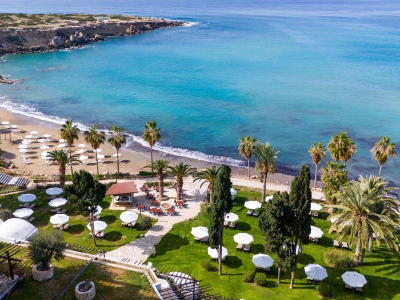Küstenlandschaft mit blauem Meer, Sandstrand, Palmen und grüner Gartenanlage mit weißen Sonnenschirmen.