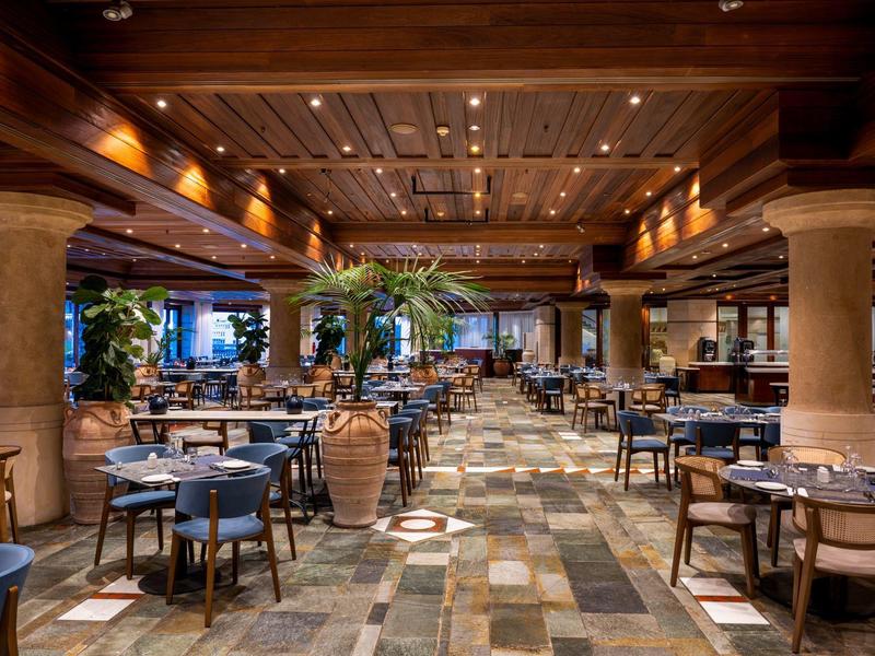 Großes Restaurant mit holzverkleidetem Decken, Marmorboden und runden Tischen mit blauen Stühlen.