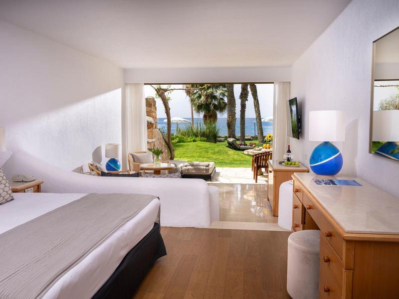 Helles Schlafzimmer mit großem Fensterblick auf Garten, Meer und Palmen, Holzfußboden, weißem Bett.