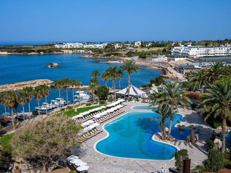 Ein großer Pool mit Liegestühlen umgeben von Palmen, Blick auf Hafen und Küstenstadt im Hintergrund.