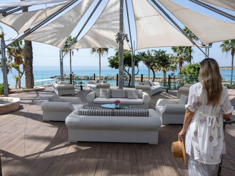 Strandcafé mit weißen Möbeln und Sonnenschirmen, Palmen und Meer im Hintergrund, Frau im Kleid.