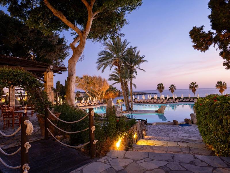 Poolbereich mit Sonnenschirmen, Palmen und beleuchteten Wegen bei Abendlicht am Meer.