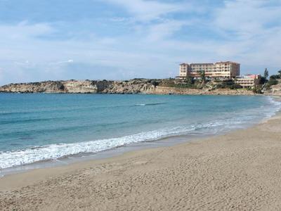Breiter Sandstrand mit ruhigem blauem Meer und Gebäuden auf felsiger Küste im Hintergrund.