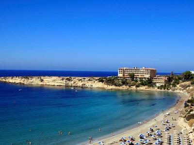 Strand mit hellem Sand, türkisblauem Wasser und klarem, blauem Himmel, Hotel im Hintergrund.