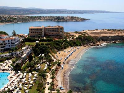 Küstenhotel mit Pool, Strand mit Liegen, hellblauem Meer und bewachsenen Hügeln im Hintergrund.