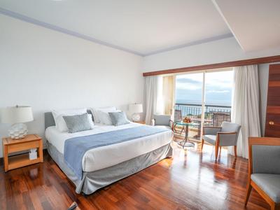 Helles Hotelzimmer mit großem Bett, Holzboden und Blick auf Meer durch große Fenster.