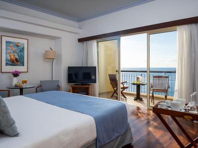Helles Hotelzimmer mit Balkon, Meerblick und gemütlicher Einrichtung mit Bett und Sitzbereich.
