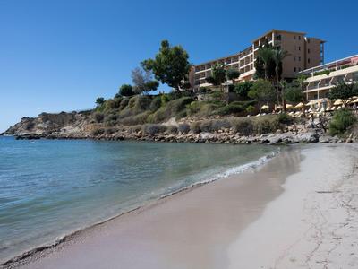Sonniger Strand mit klarem Wasser und Hotels auf einer felsigen Anhöhe am Ufer.