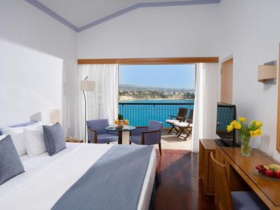 Helles Hotelzimmer mit Meerblick, großem Bett, Schreibtisch und Balkon mit Sitzgelegenheiten.