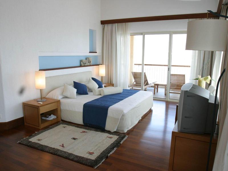 Helles Schlafzimmer mit Holzboden, Doppelbett, Nachttisch, Teppich und Balkon mit Aussicht.