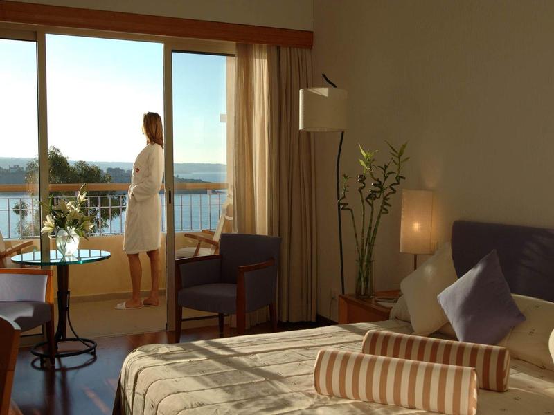 Gemütliches Schlafzimmer mit Bett, Nachttischlampen und Frau am Balkon mit Meerblick.