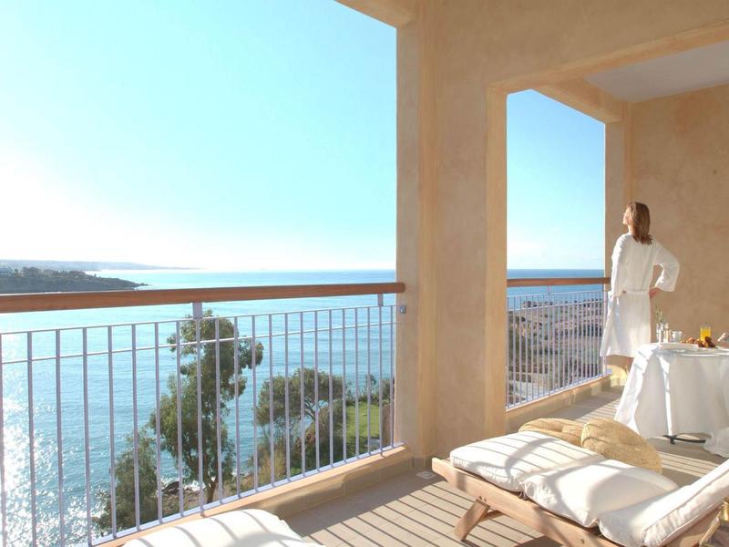 Balkon mit Liegestuhl, Tisch und Blick auf das blaue Meer bei klarem Himmel.