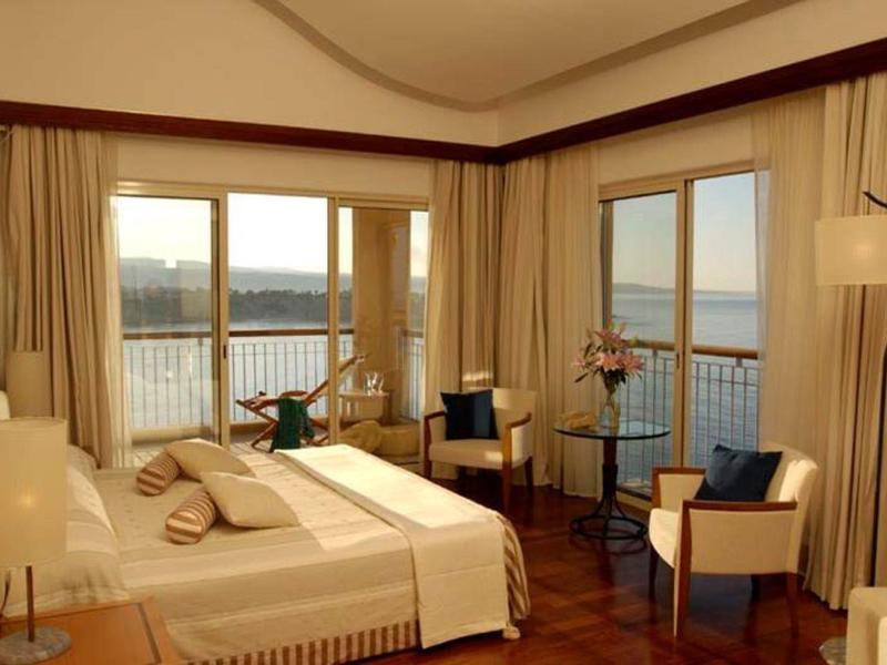 Hell eingerichtetes Schlafzimmer mit Balkon, Doppelbett, zwei Stühlen und weitem Blick aufs Wasser.