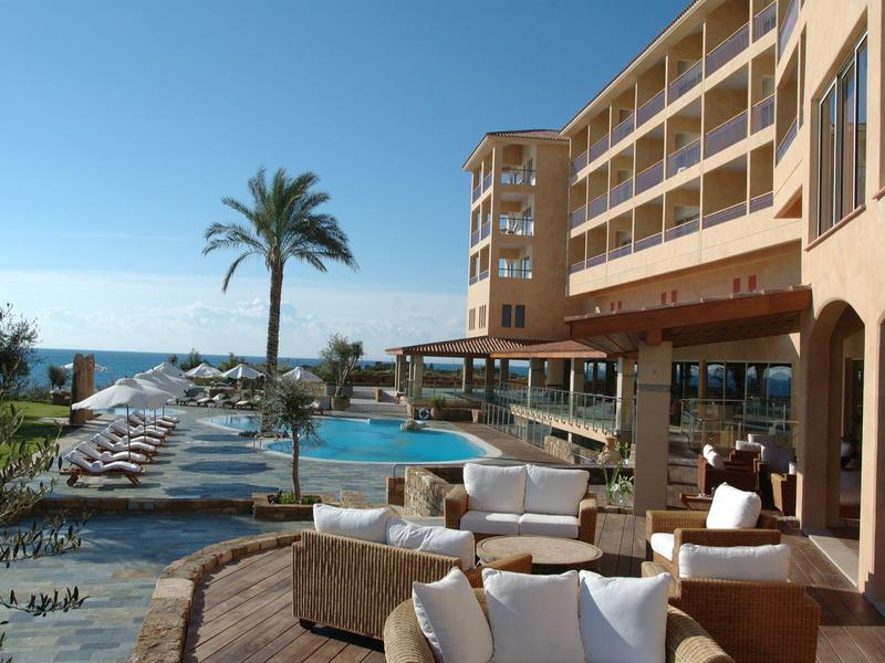 Hotel mit Terrasse, Pool, Palmen und Blick auf das Meer bei klarem Himmel.