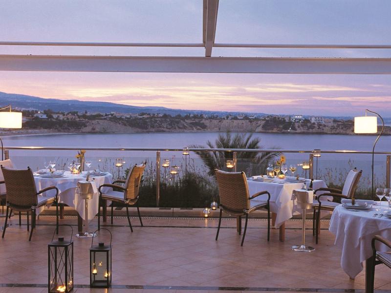 Offenes Terrassenrestaurant mit weißen Tischtüchern, Blick auf Wasser und Berge im Abendlicht.