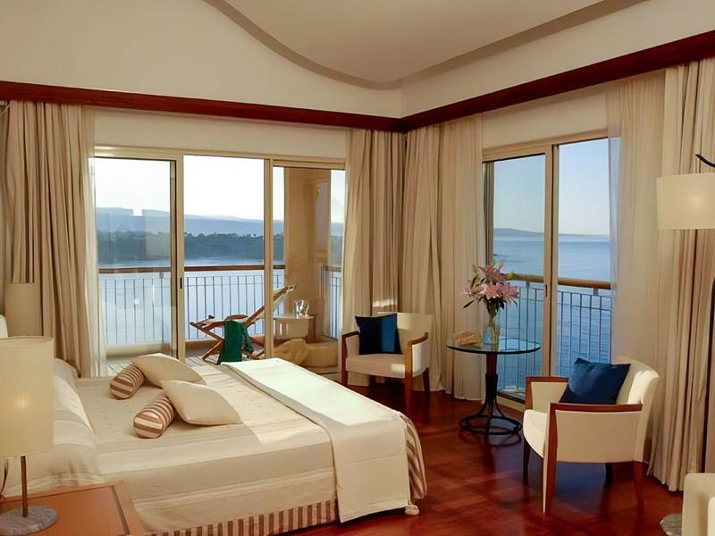 Helles Schlafzimmer mit großen Fenstern, Holzfußboden, Bett, Sesseln und Meerblick.