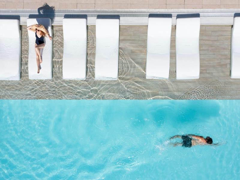 Blick von oben auf einen Pool mit blauem Wasser, einer schwimmenden Person und einer liegenden Person auf Liegestühlen.