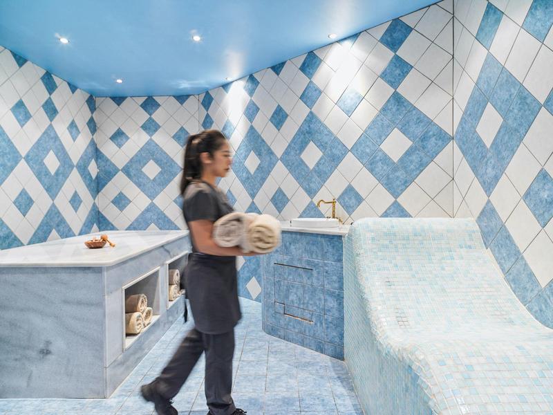 Frau in modernem Dampfbad mit blau-weißen Fliesen und gerollten Handtüchern in hellem Raum.