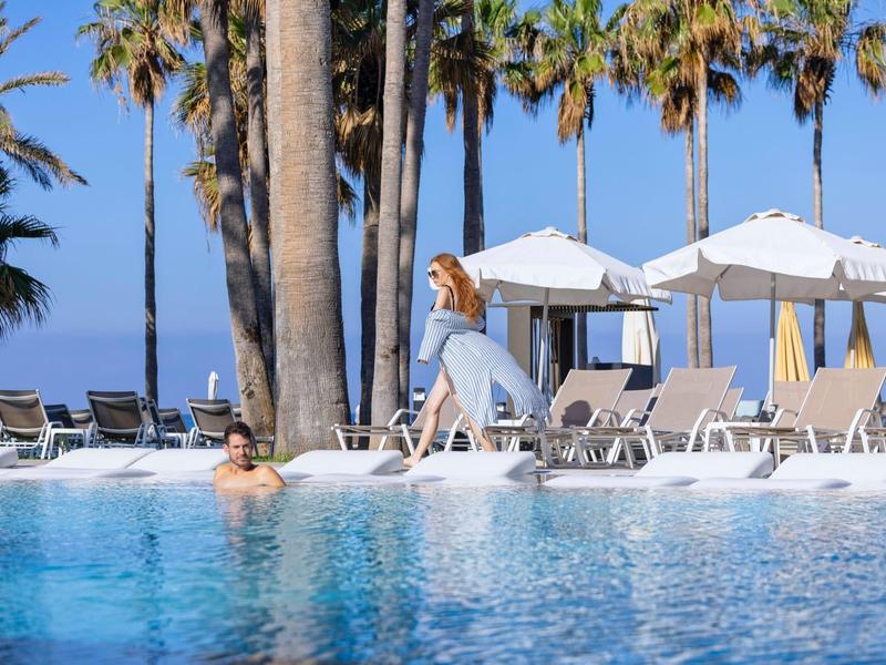 Schwimmbecken mit blauer Wasseroberfläche, Liegestühlen, Palmen und einer Frau mit Hut am Rand.