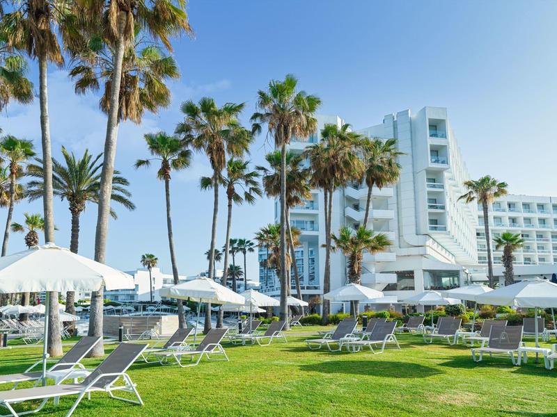Grüner Garten mit Liegestühlen und weißen Sonnenschirmen vor Palmen und weißem Hotelgebäude.