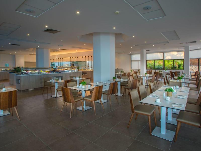 Moderne Cafeteria mit Holzstühlen, weißen Tischen und großen Fenstern, heller Raum mit Deckenspots.