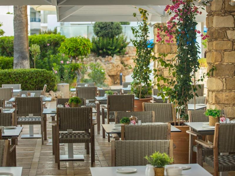 Außenbereich eines Cafés mit vielen Holztischen und Stühlen, grünen Pflanzen und Steinwänden.