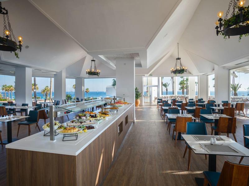 Großes, helles Restaurant mit Blick aufs Meer, modernen Tischen, Stühlen und Buffetstation in der Mitte.