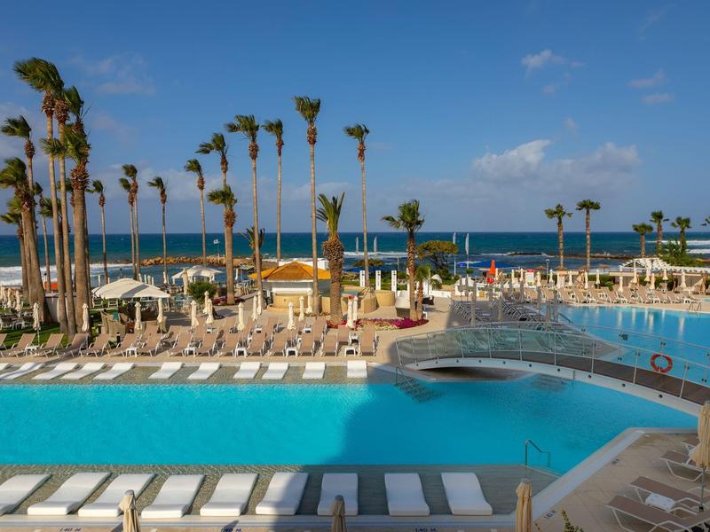 Großer Pool mit weißem Liegestuhl, Palmen und Meer unter blauem Himmel im Hintergrund.