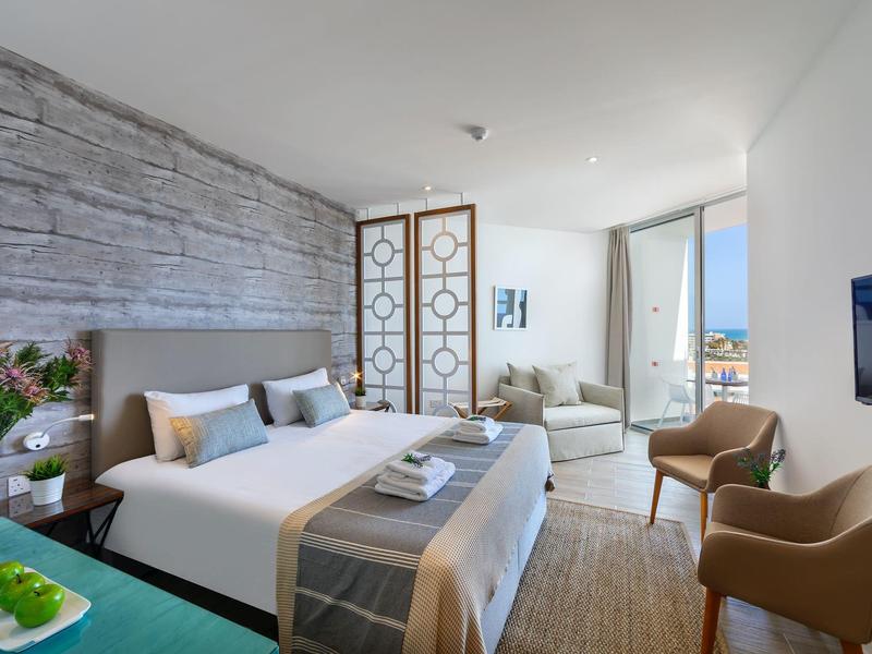 Hell eingerichtetes Schlafzimmer mit Doppelbett, Sitzbereich, braunen Sesseln und Fenster mit Meerblick.