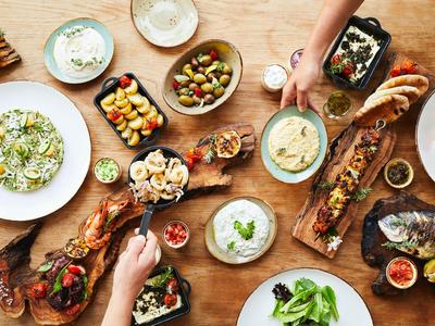 Tisch mit verschiedenen mediterranen Gerichten, Brot und Dips, Hände greifen nach Essen.