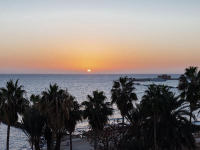 Sonnenuntergang über dem Meer mit Palmen im Vordergrund an einem Strandhotel.