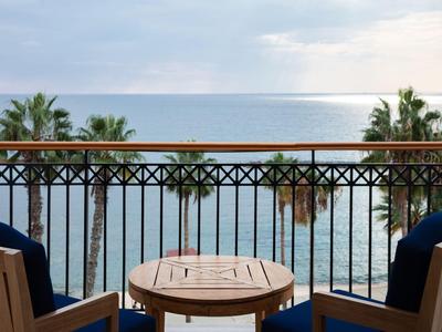 Balkon mit Holztisch und Stühlen, Blick auf Meer und Palmen bei bewölktem Himmel.