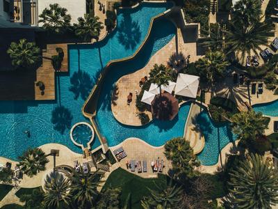 Luftaufnahme eines geschwungenen Hotelpools mit umliegenden Liegestühlen und Sonnenschirmen.