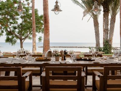 Freiluftrestaurant mit Holztischen und -stühlen, Blick aufs Meer und Palmen.