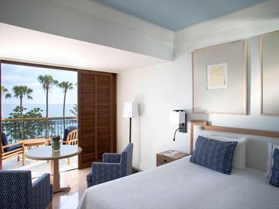 Helles Hotelzimmer mit Doppelbett, Sitzbereich und Balkon mit Meerblick und Palmen.