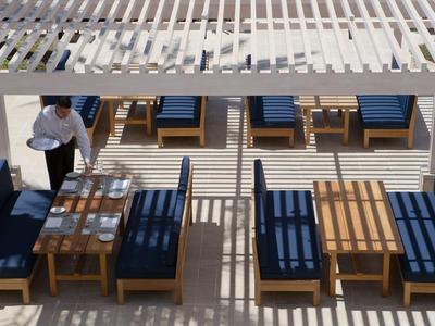 Servierpersonal deckt draußen an Tischen mit blauen Polstern unter einer Sonnenterrasse ein.