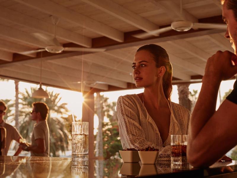 Menschen sitzen in einem hellen, offenen Café mit Holzdach bei Sonnenuntergang.