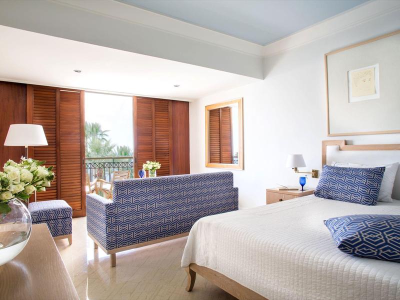 Helles Schlafzimmer mit weißen Wänden, blauem Sofa, Doppelbett und großen Holzfenstern mit Balkon.