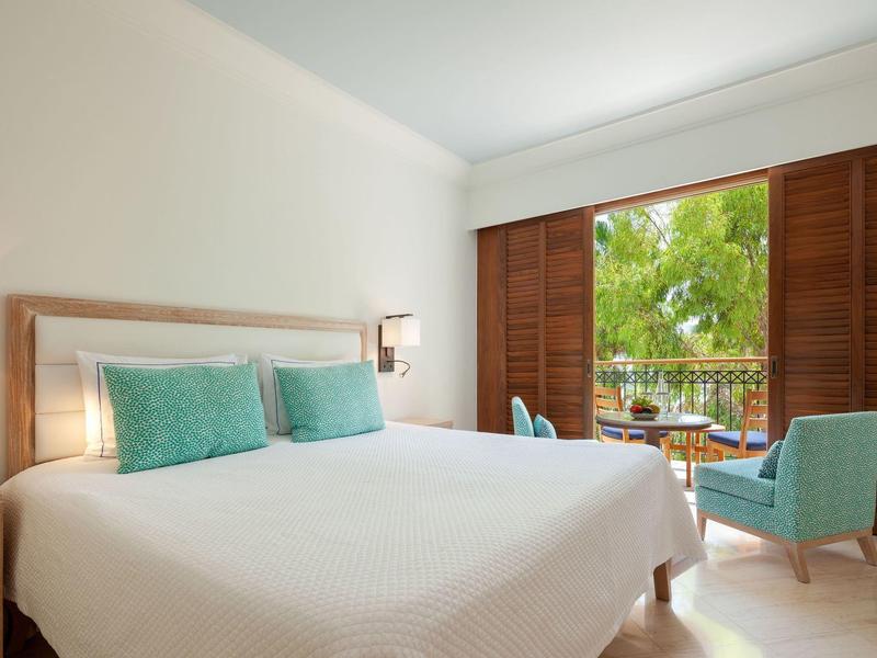 Helles Schlafzimmer mit großem Bett, türkisfarbenen Kissen und Stühlen, und offenem Fenster zur Terrasse.