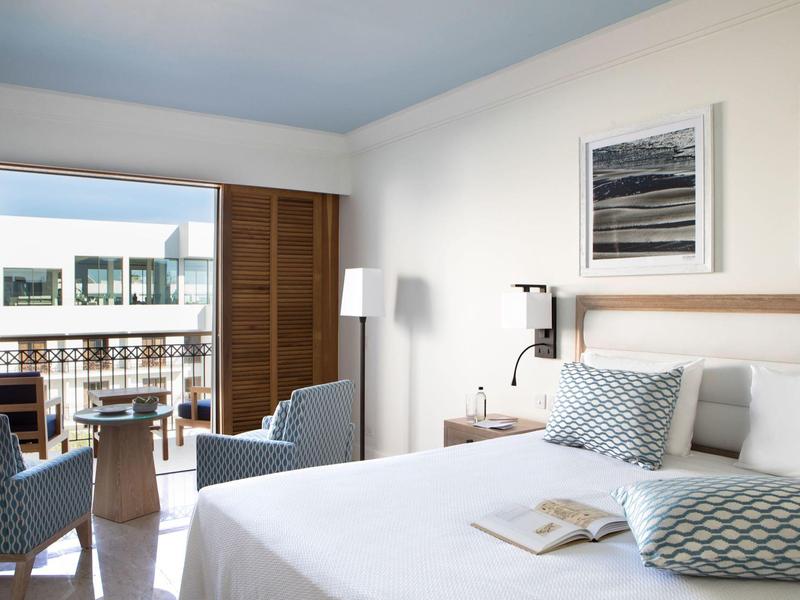 Hell eingerichtetes Hotelzimmer mit Doppelbett, Stühlen, Tisch und Balkon mit Blick nach draußen.