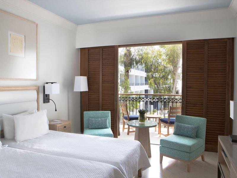 Helles Hotelzimmer mit Doppelbett, zwei hellblauen Sesseln und Balkon mit Holztüren.