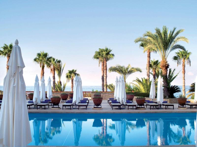 Ein klarer Pool mit blauen Liegen und weißen Sonnenschirmen, umgeben von Palmen und blauem Himmel.