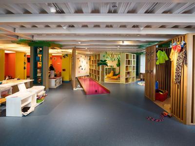 Großer heller Raum mit Holzmöbeln, bunten Sitzflächen und einer blauen Bodenfläche.